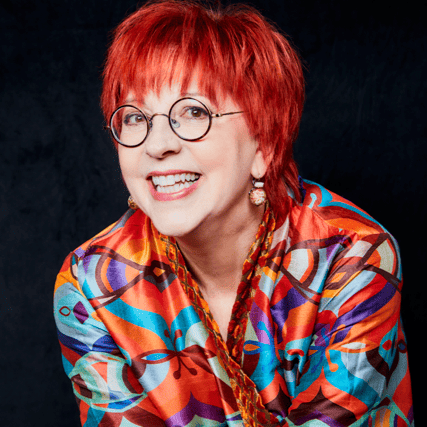 Une personne souriante aux cheveux courts et d'un rouge éclatant porte des lunettes rondes et une veste à motifs colorés sur un fond sombre.