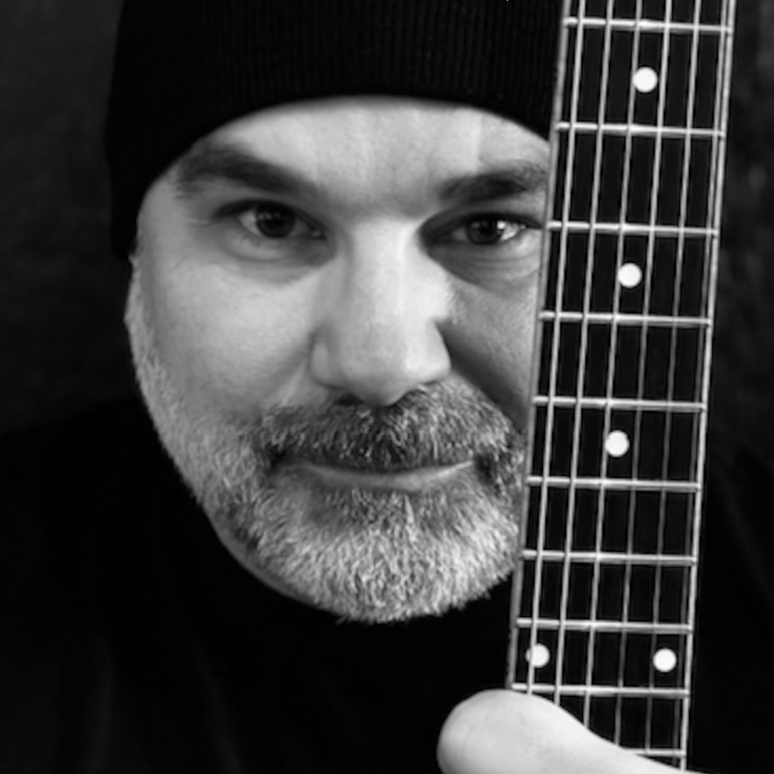 Photo en noir et blanc d'un homme portant un bonnet, une barbe et une moustache, tenant un manche de guitare verticalement devant son visage, couvrant partiellement un œil.
