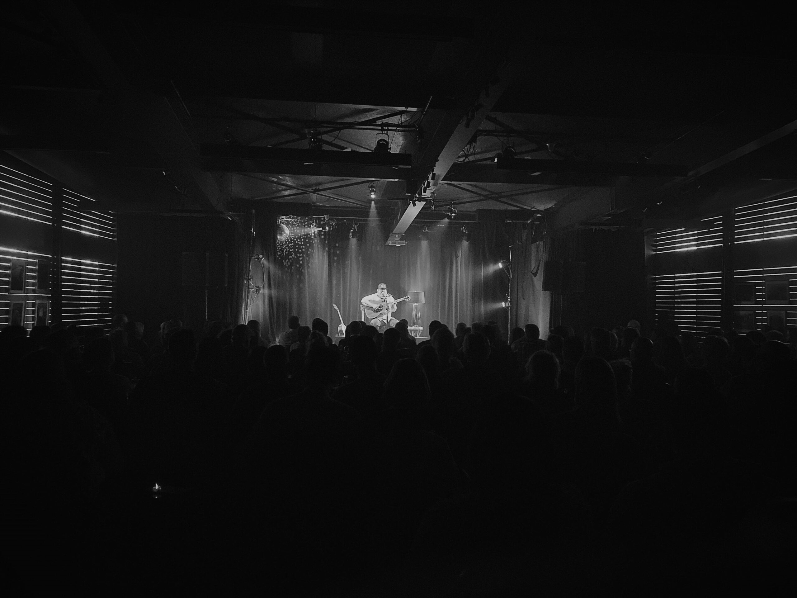Un musicien est assis sur scène et joue de la guitare acoustique dans une salle faiblement éclairée par des projecteurs, tandis qu'un public le regarde au premier plan. L'atmosphère est intime et l'image est en noir et blanc.