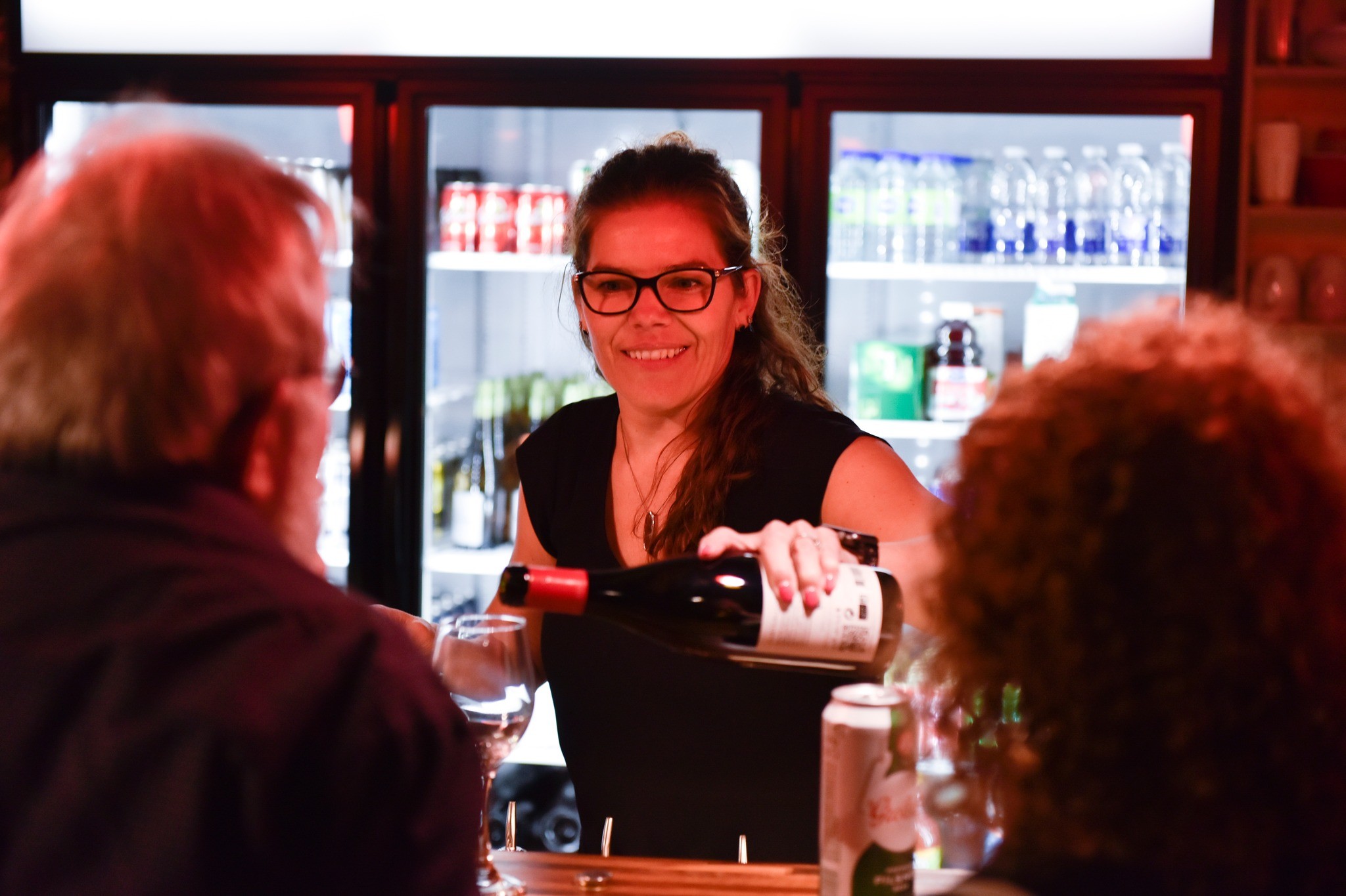 Un barman souriant avec des lunettes verse du vin rouge dans un verre pour deux clients à un bar, avec des réfrigérateurs remplis de boissons visibles à l'arrière-plan.