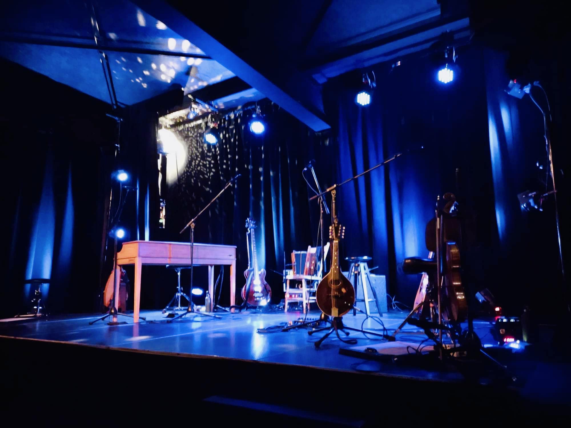 Une scène faiblement éclairée par des projecteurs bleus met en lumière divers instruments de musique, dont un clavier, des guitares et un violoncelle, équipés de microphones et de pieds, prêts à être joués en direct.