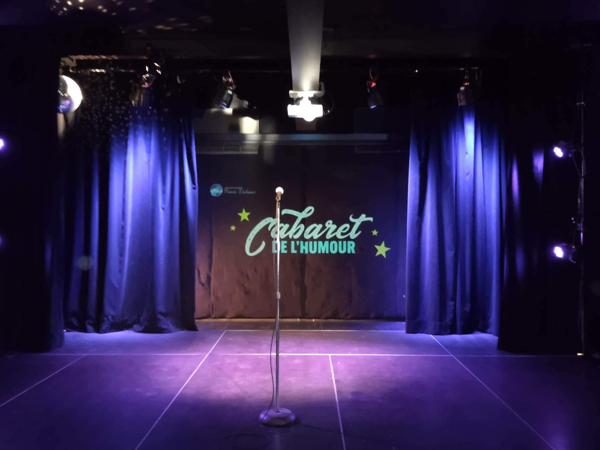 Un projecteur éclaire le pied d'un microphone au centre de la scène, tandis que des rideaux sombres et un panneau indiquant "Cabaret de l'Humour" sont éclairés en arrière-plan. L'atmosphère suggère une salle de comédie ou de spectacle.