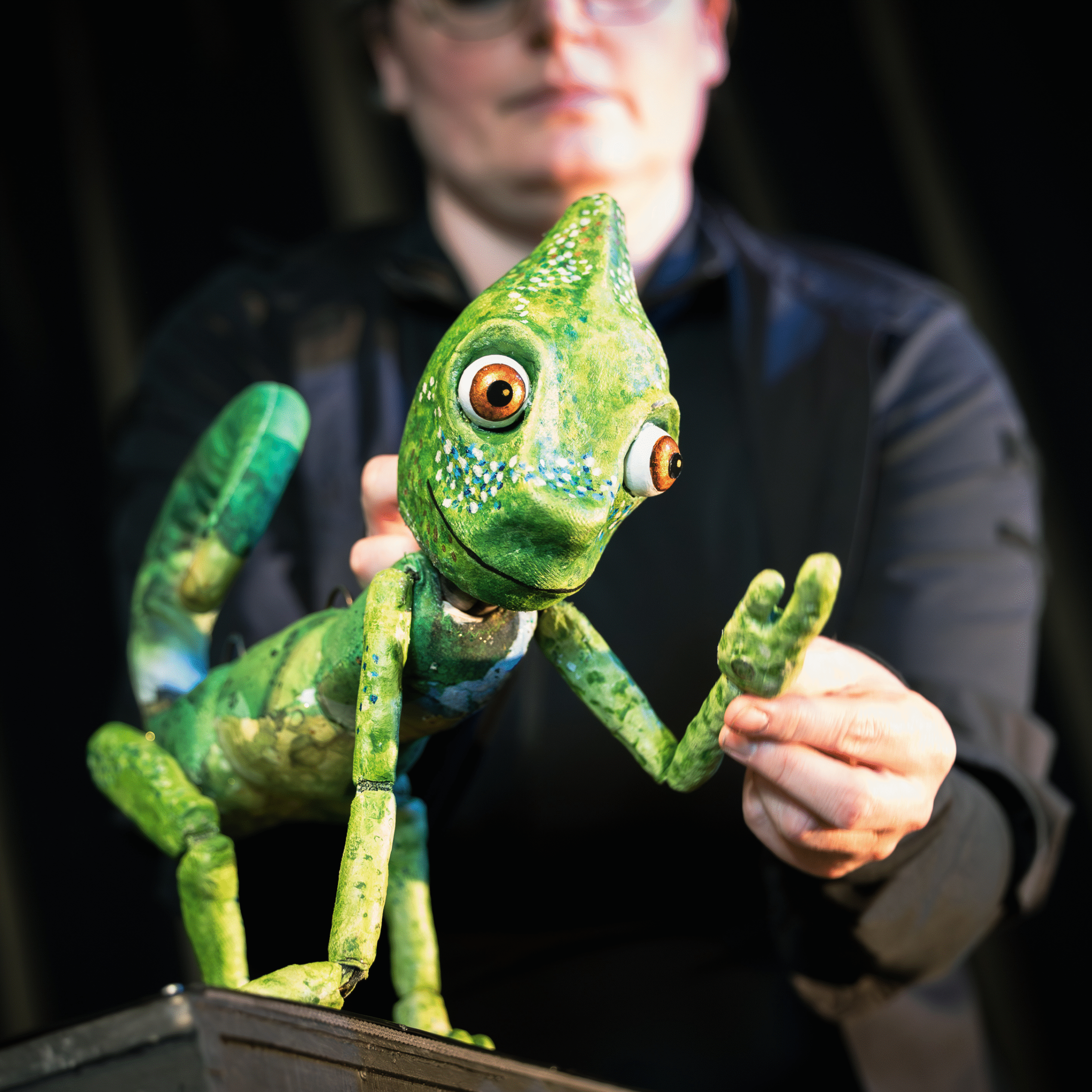Une personne manipule une marionnette caméléon verte colorée avec de grands yeux, en levant le bras de la marionnette comme si elle faisait un signe de la main. L'accent est mis sur la marionnette, le marionnettiste étant flou à l'arrière-plan.