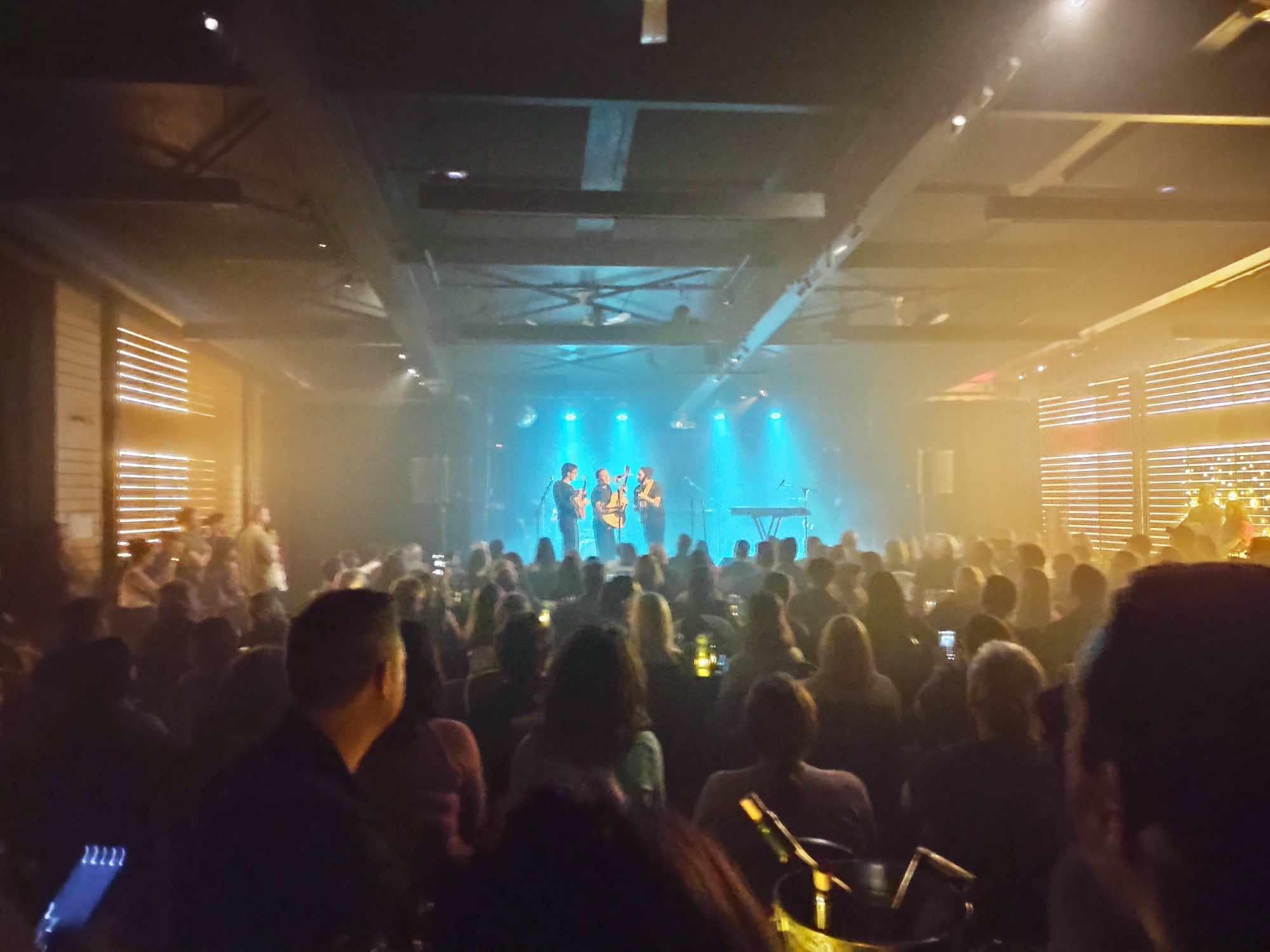 Un public nombreux regarde un groupe se produire sur scène dans une salle intérieure faiblement éclairée, avec un éclairage de scène bleu et des personnes assises et debout dans toute la salle.