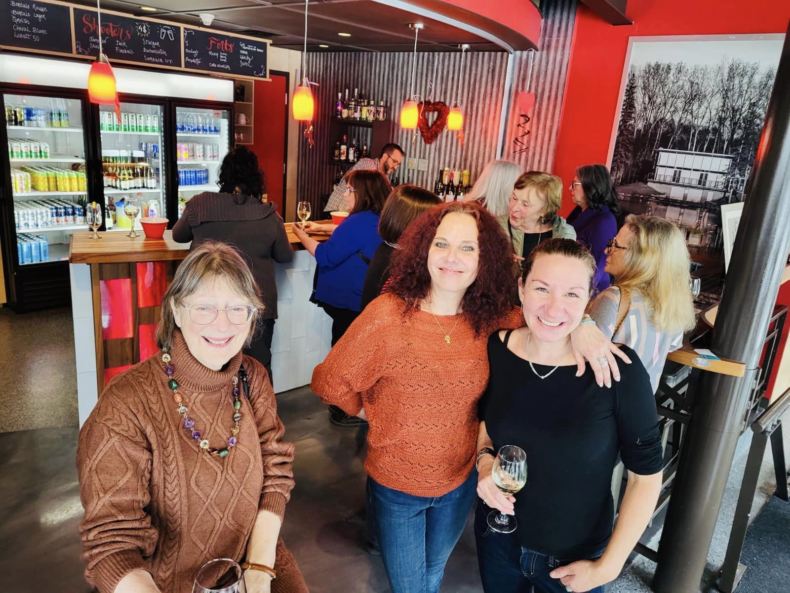 Un groupe de femmes, dont certaines tiennent un verre et sourient, se réunit dans un bar confortable aux murs rouges, à la devanture en tôle ondulée et au tableau des menus situé au-dessus de réfrigérateurs remplis de boissons. Une photo en noir et blanc orne le mur.