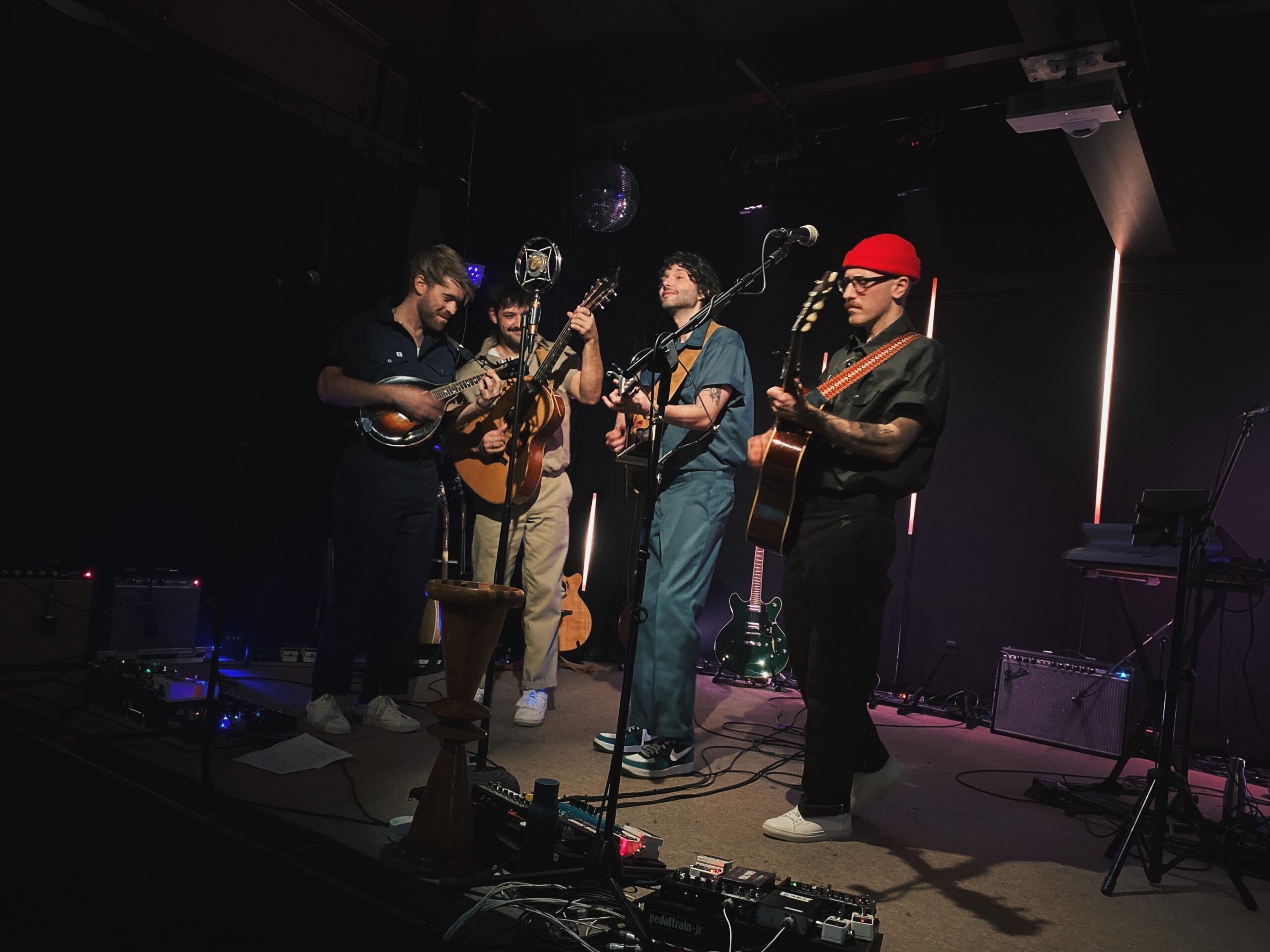 Quatre musiciens se tiennent près les uns des autres sur une scène faiblement éclairée, chacun jouant d'un instrument à cordes, notamment d'une guitare et d'une mandoline. Ils semblent jouer dans une salle intime, avec des équipements et des câbles visibles autour d'eux.