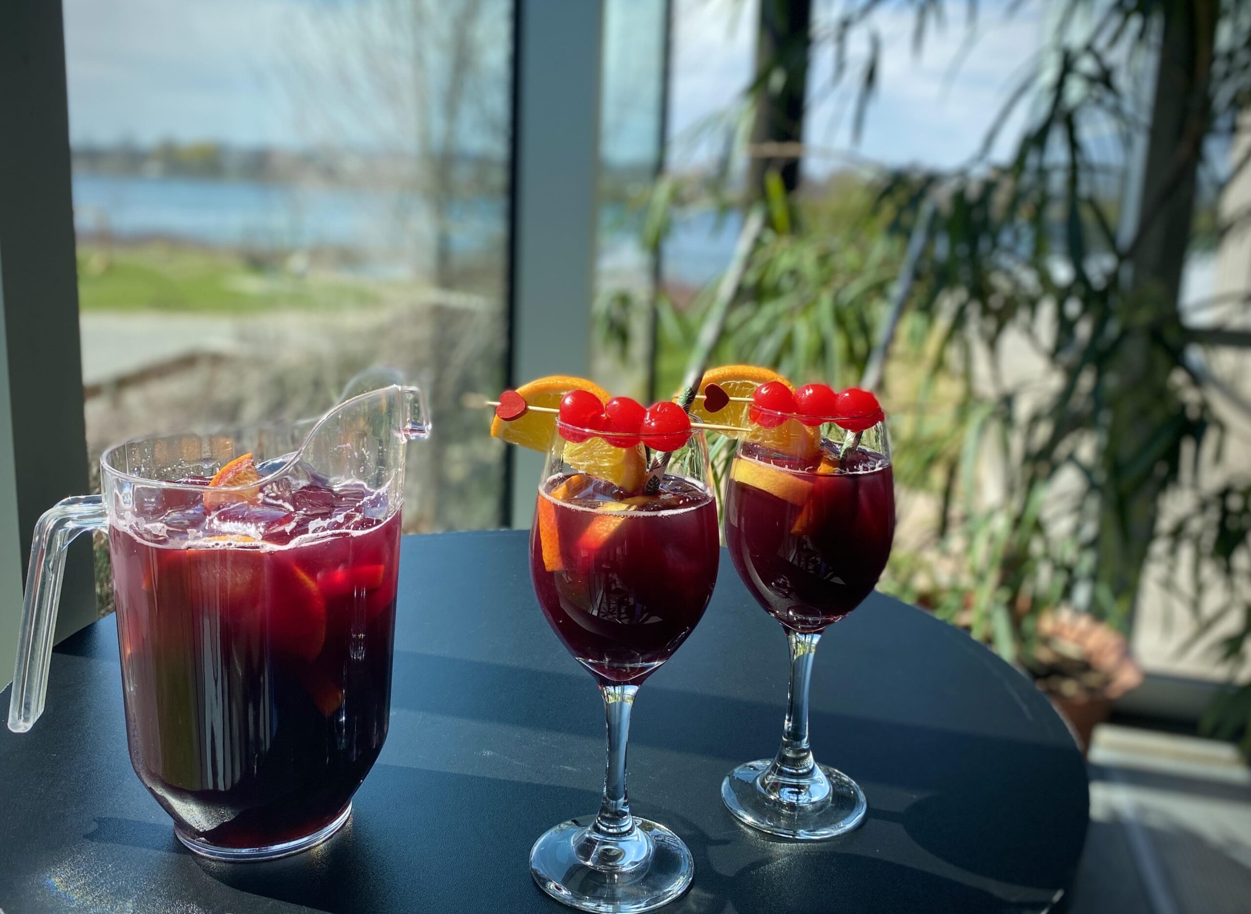 Un pichet et deux verres de sangria rouge avec des glaçons, des tranches d'orange et des cerises sont posés sur une table près d'une fenêtre ensoleillée qui offre une vue panoramique sur l'extérieur.