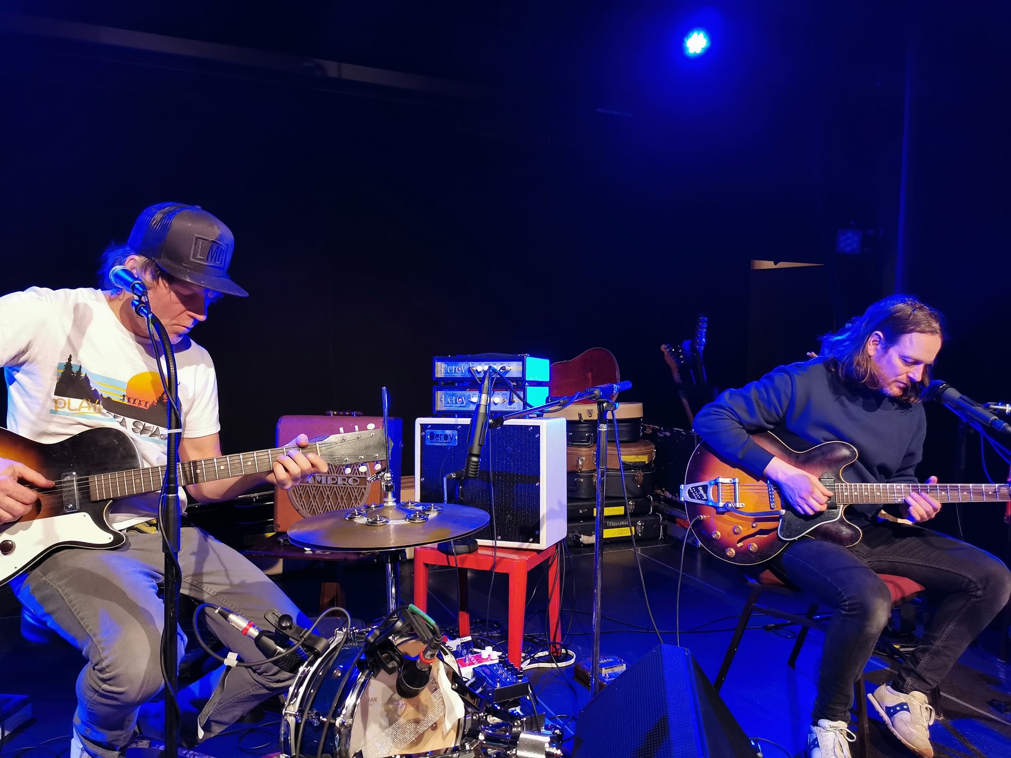 Deux musiciens sont assis sur scène et jouent de la guitare électrique, entourés d'amplificateurs, de microphones et de divers équipements musicaux. L'un porte une chemise blanche et une casquette, l'autre un sweat à capuche sombre. L'éclairage bleu de la scène met en valeur la scène.