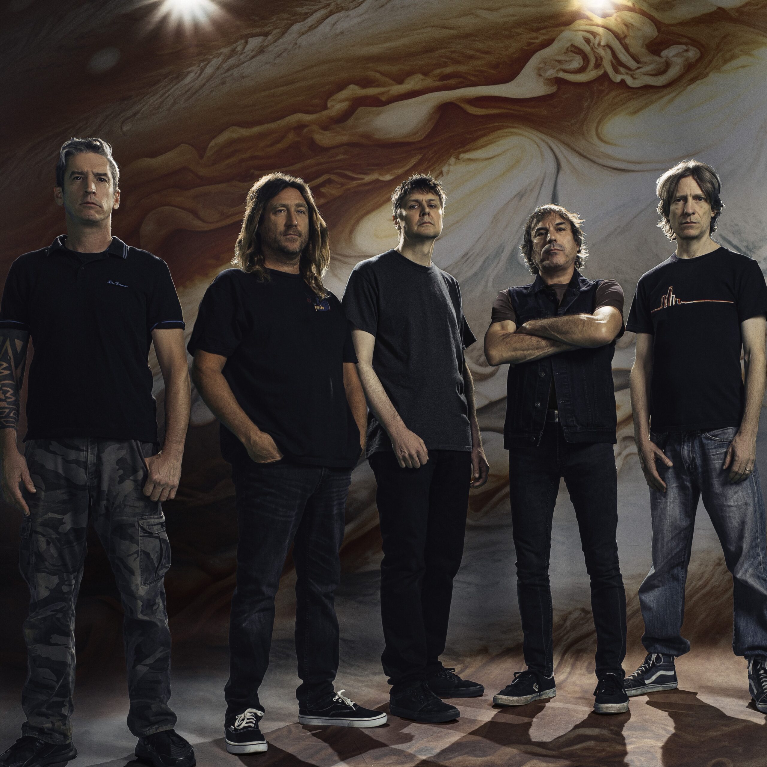 Cinq hommes se tiennent côte à côte sur une toile de fond tourbillonnante d'inspiration spatiale. Ils portent des vêtements sombres et décontractés et regardent sérieusement l'appareil photo, les bras le long du corps ou croisés.