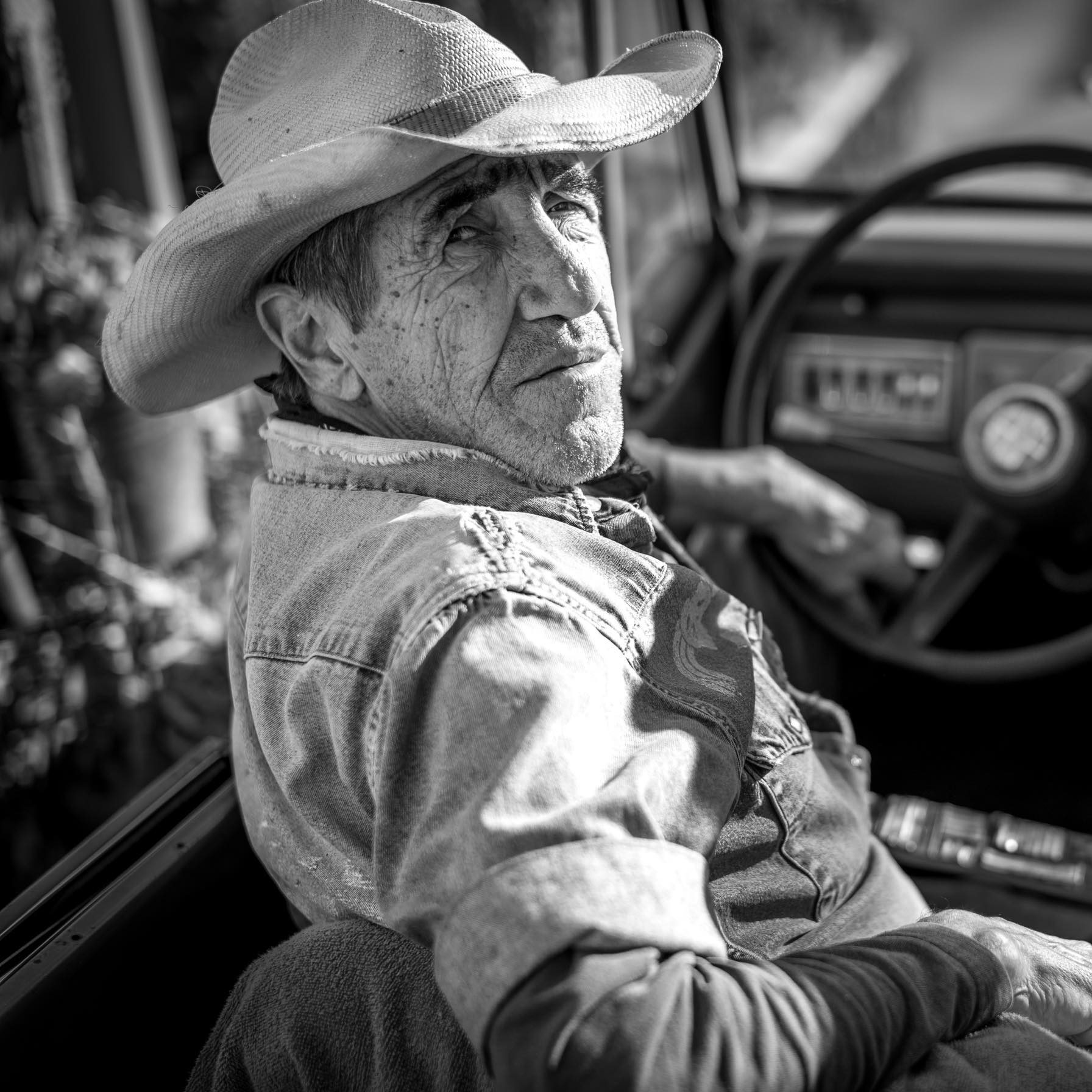 Un homme âgé portant un chapeau de cow-boy et une veste en jean est assis sur le siège du conducteur d'un véhicule et regarde par-dessus son épaule en direction de l'appareil photo. L'image est en noir et blanc, la lumière du soleil éclairant son visage.