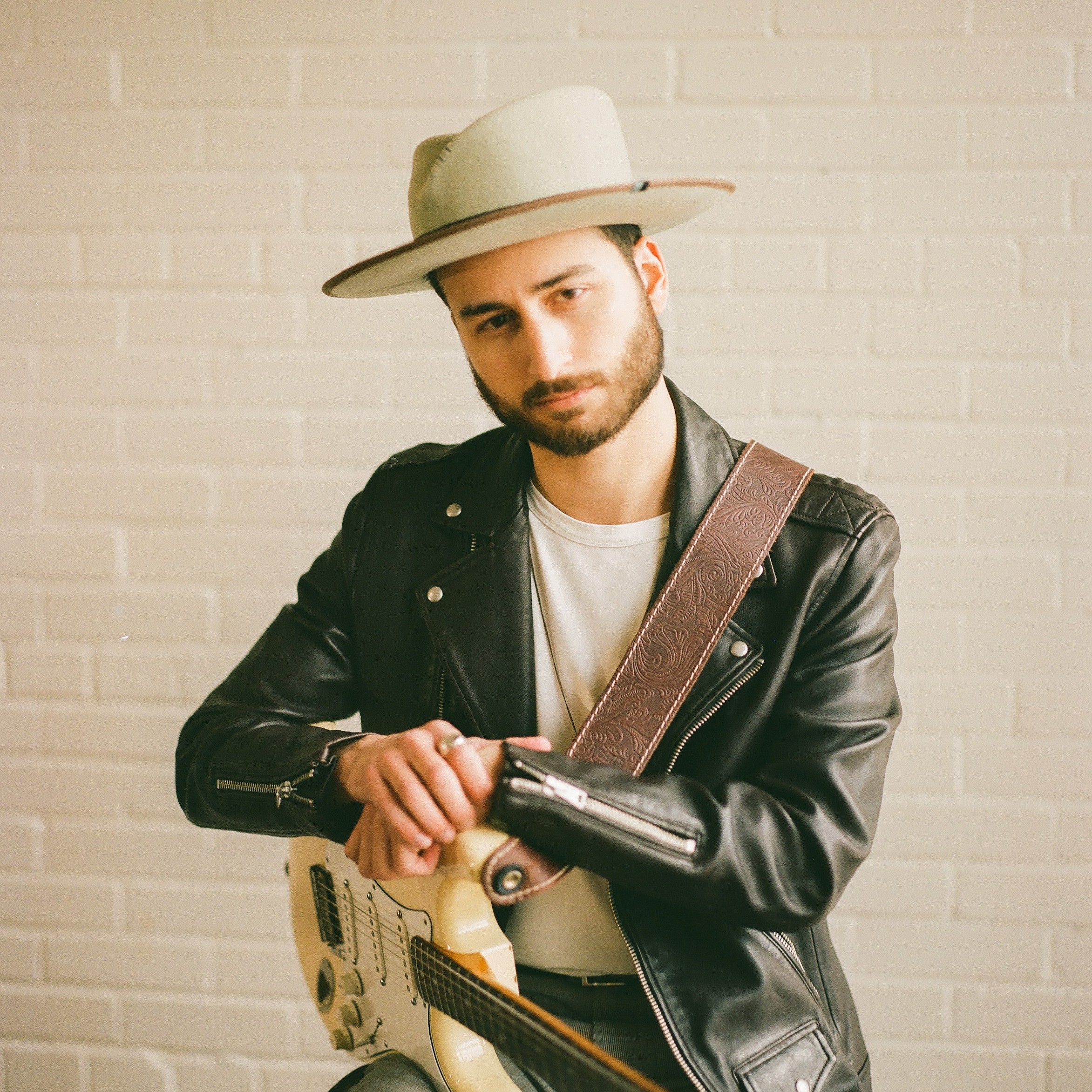 Un homme portant un chapeau à larges bords, une veste en cuir noir et une chemise blanche tient une guitare électrique et regarde la caméra, debout devant un mur de briques blanches.