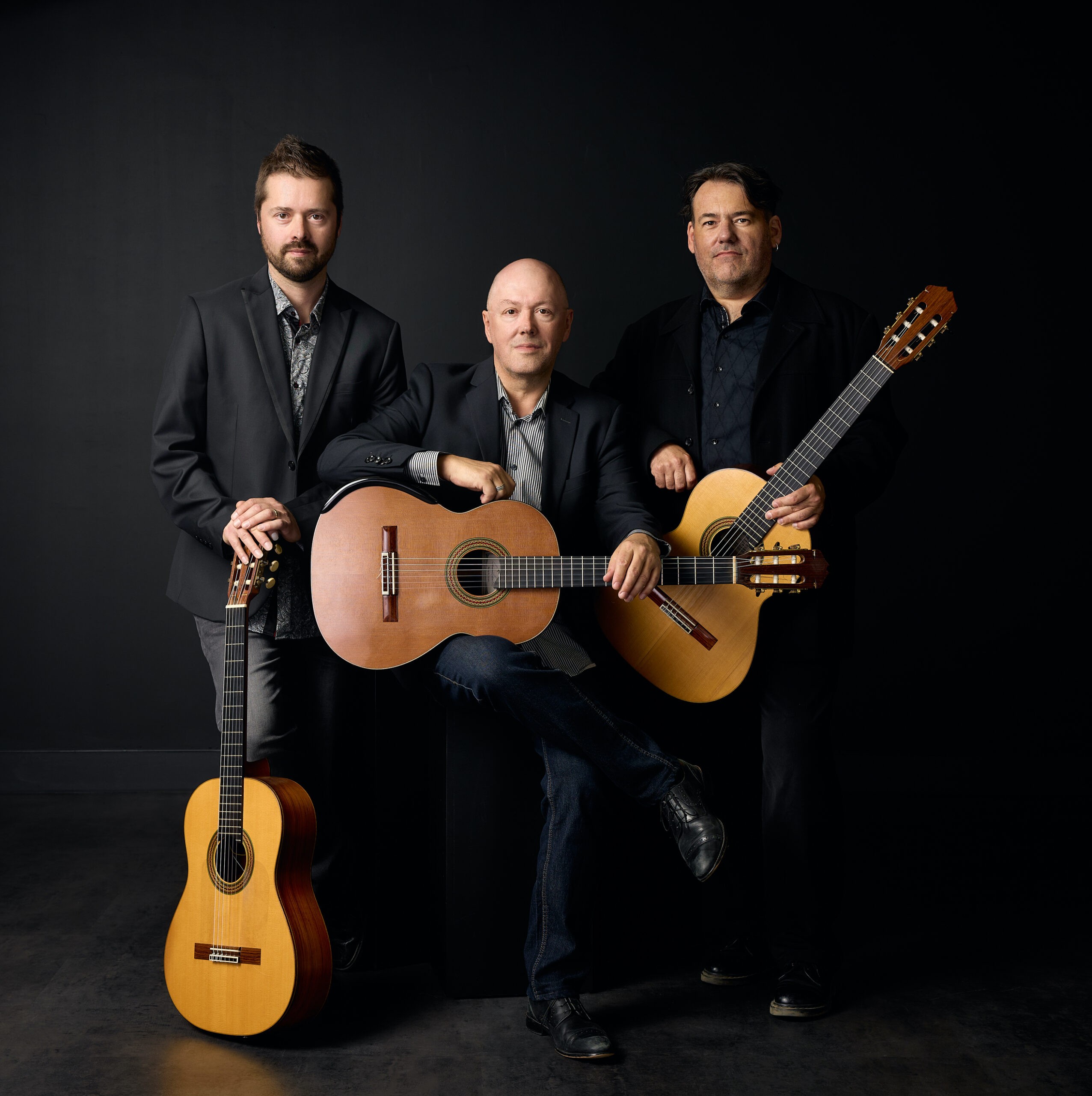 Trois hommes vêtus de costumes sombres posent avec des guitares classiques sur un fond sombre. Deux d'entre eux se tiennent debout de chaque côté, les guitares à la main, tandis que l'homme au centre est assis avec sa guitare sur les genoux, tous regardant l'appareil photo.