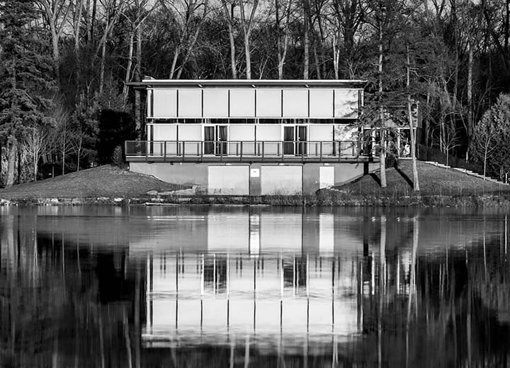 Un bâtiment moderne, rectangulaire et doté de grandes fenêtres, se trouve au bord d'un lac calme, se reflétant clairement dans l'eau. Des arbres sans feuilles entourent la structure et la scène est en noir et blanc.
