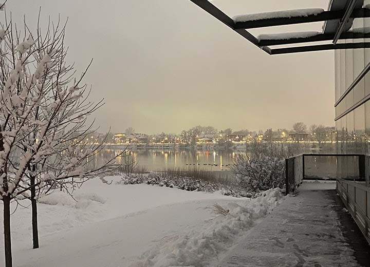Un paysage enneigé avec un arbre et un sentier à côté d'un bâtiment, surplombant une rivière calme. Des maisons illuminées sont visibles de l'autre côté de l'eau, sous un ciel nuageux et hivernal.