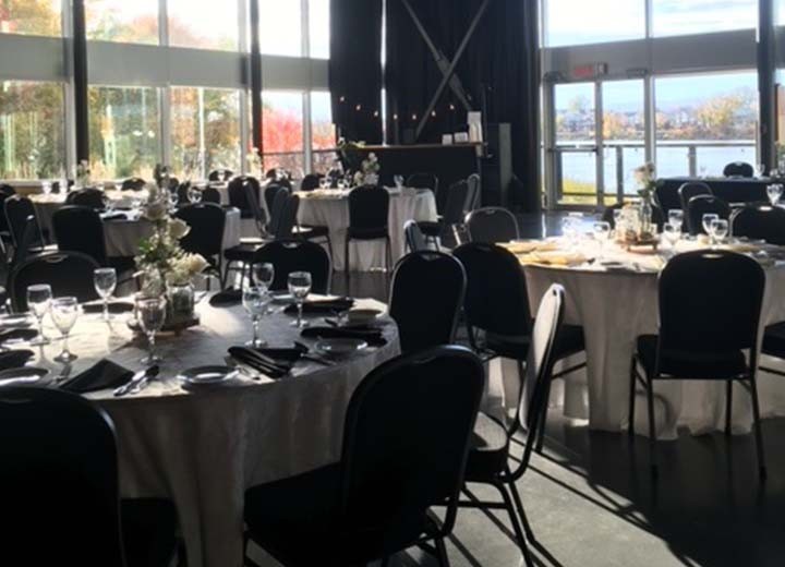 Salle de banquet ensoleillée avec des tables rondes préparées pour un événement, décorées de nappes blanches, de verres et de centres de table floraux. De grandes fenêtres révèlent une vue extérieure avec des arbres et de l'eau.