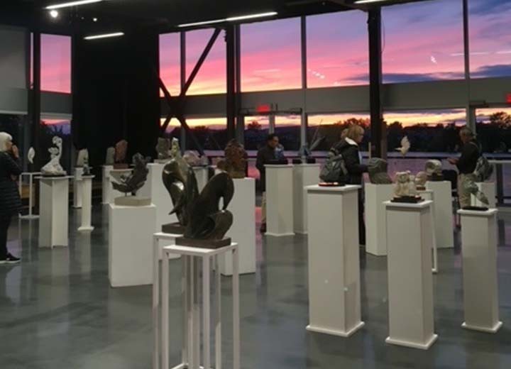 Des personnes observent diverses sculptures exposées sur des socles blancs dans une galerie moderne dotée de grandes fenêtres. Un ciel coloré au coucher du soleil est visible à l'extérieur et projette de la lumière dans la pièce.