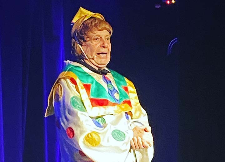 Une personne déguisée en clown dans un costume coloré avec un chapeau pointu et des pois, se produisant sur scène sous des lumières vives.