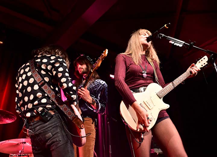 Trois musiciens se produisent sur scène sous un éclairage rouge. Une femme vêtue d'une tenue rouge chante dans un micro tout en jouant de la guitare électrique, tandis que deux hommes jouent de la guitare à ses côtés.