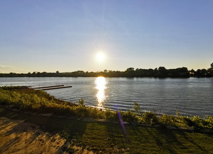 Le soleil se couche sur une rivière calme, jetant un reflet lumineux sur l'eau. Des arbres bordent la rive opposée et un petit quai s'avance dans la rivière près d'une rive herbeuse et ombragée. Le ciel est clair et bleu.