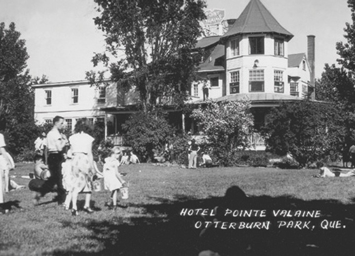 Photo en noir et blanc de personnes rassemblées sur une pelouse devant un grand bâtiment de style victorien avec une tourelle. Le texte se lit comme suit : "Hôtel Pointe Valaine, Otterburn Park, Québec. Des arbres entourent la scène.