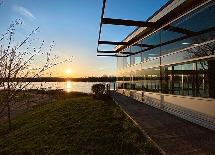 Un bâtiment moderne doté de grandes baies vitrées surplombe une rivière calme au coucher du soleil, le soleil étant bas dans le ciel et les arbres se profilant à l'arrière-plan. Une pelouse et un arbre dénudé se trouvent au premier plan.