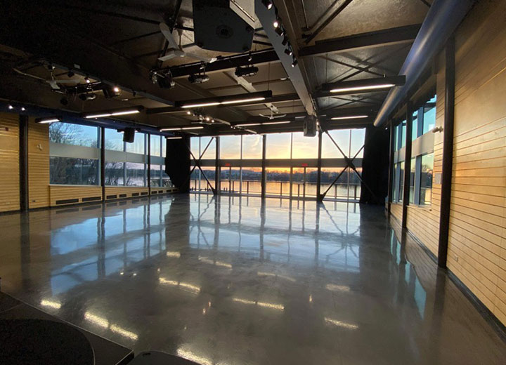 Une pièce spacieuse et vide avec un sol brillant, des murs en bois, un plafond aux poutres apparentes et de grandes fenêtres du sol au plafond montrant un coucher de soleil sur une étendue d'eau.