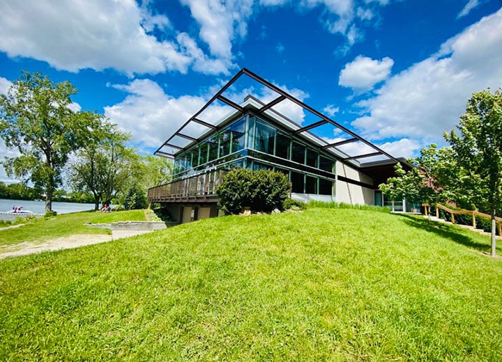 Un bâtiment moderne en verre avec un toit géométrique se trouve sur une colline herbeuse sous un ciel bleu vif avec des nuages épars. Des arbres et une rivière sont visibles à l'arrière-plan.