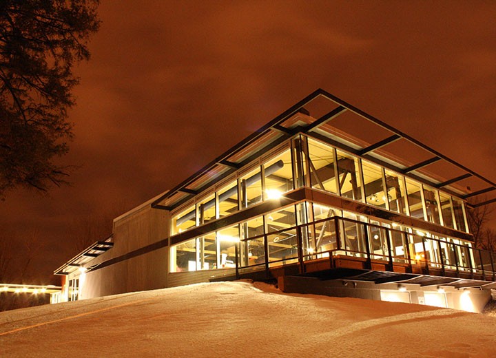 Un bâtiment moderne de verre et d'acier s'illumine de lumières chaudes la nuit, situé sur une colline enneigée sous un ciel nuageux aux teintes orangées.
