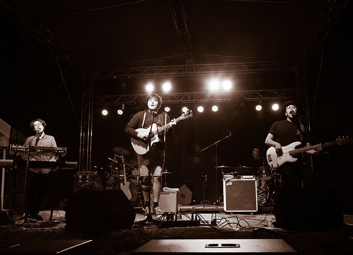 Photo en tons sépia d'un groupe se produisant sur scène la nuit. Quatre musiciens jouent du clavier, de la guitare et de la basse, avec un batteur à l'arrière. Les lumières de la scène brillent au-dessus de la tête, éclairant le groupe.