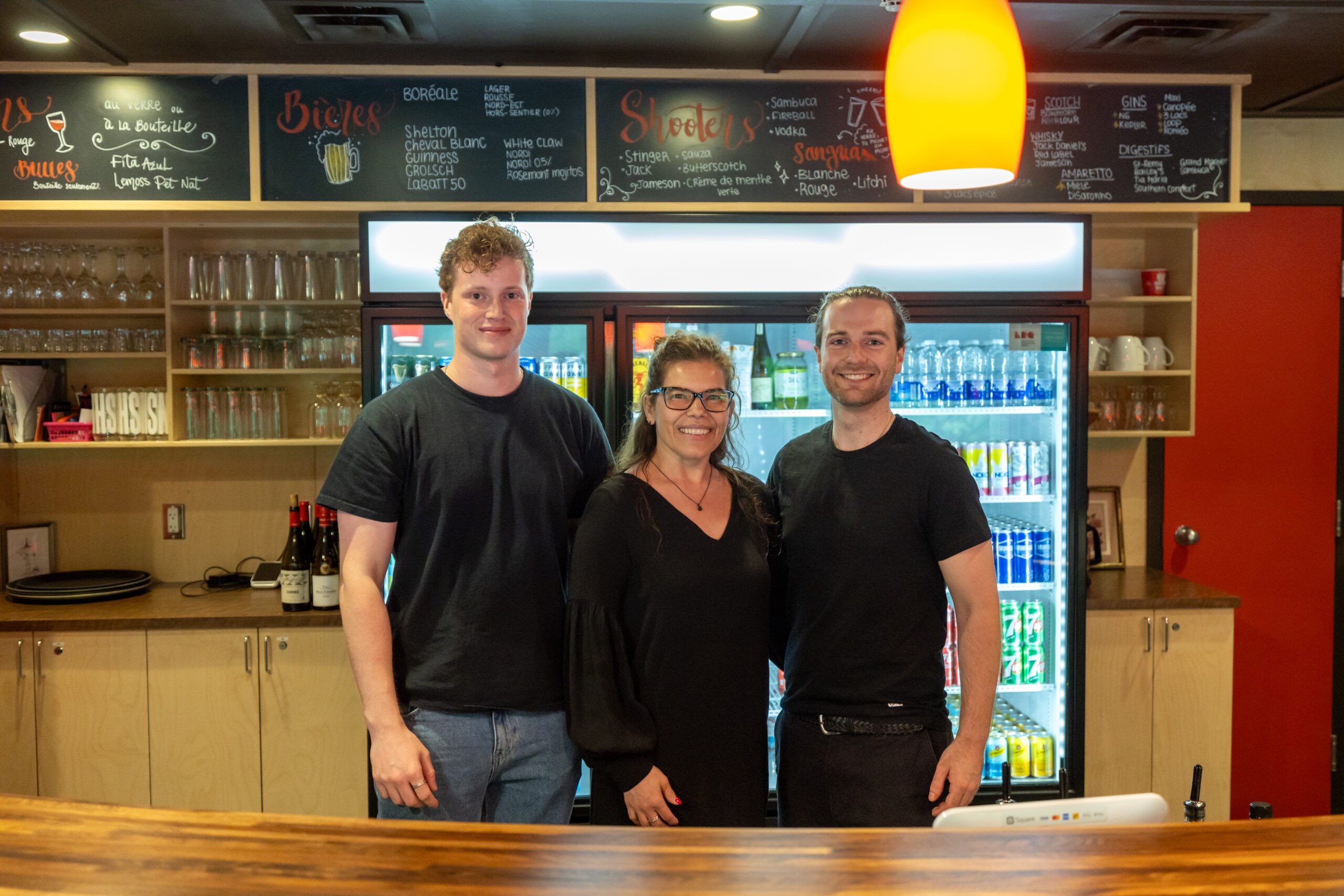 Trois personnes sourient derrière un comptoir en bois dans un café ou un restaurant, avec des étagères, un réfrigérateur rempli de boissons et un menu sur tableau en arrière-plan. Un éclairage chaud est suspendu au-dessus de la tête.