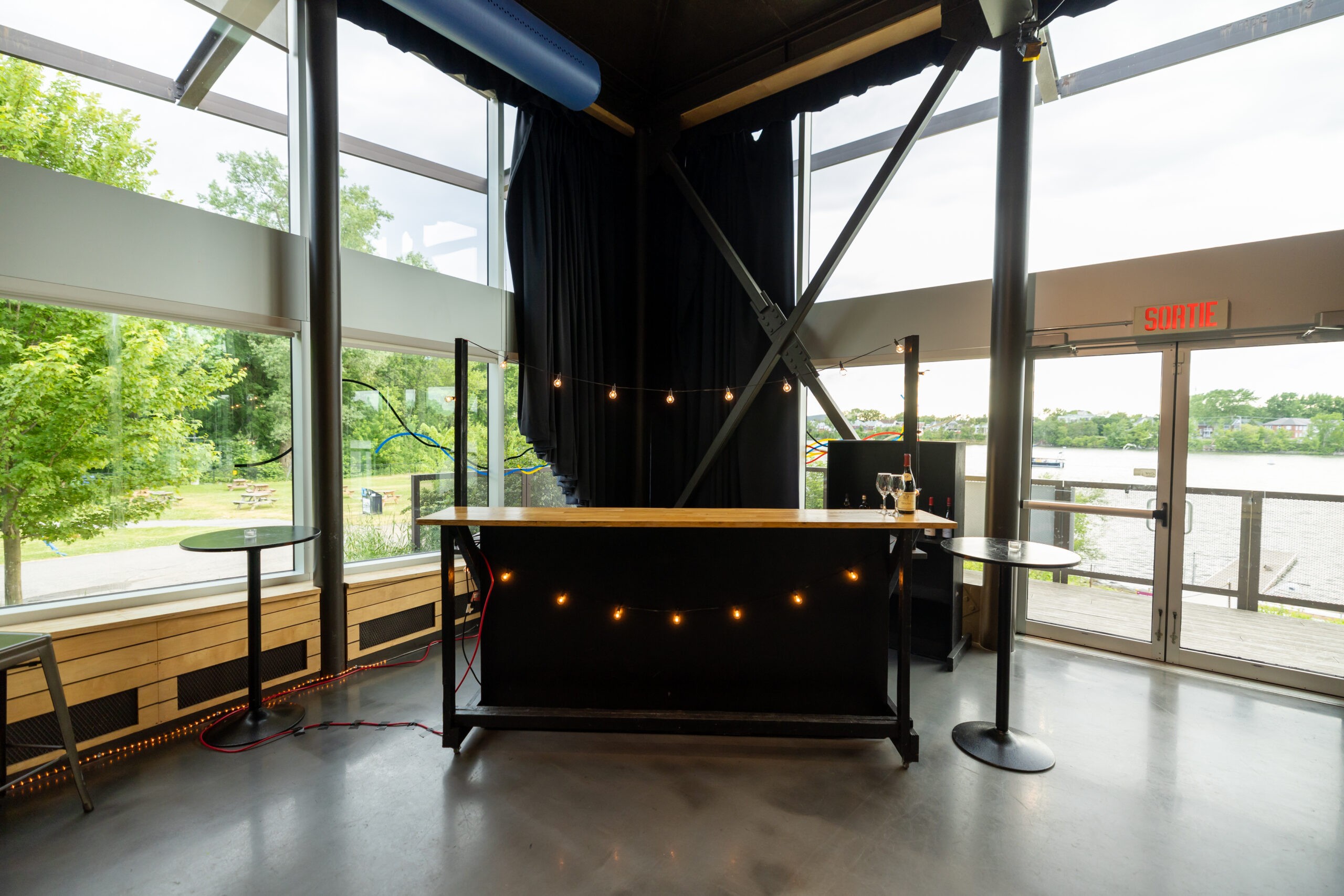 Un bar intérieur moderne avec des guirlandes lumineuses sur le comptoir, entouré de grandes fenêtres offrant une vue sur la rivière avec des arbres et des tables de pique-nique à l'extérieur. L'espace est doté de sols en béton poli et d'un panneau EXIT visible.
