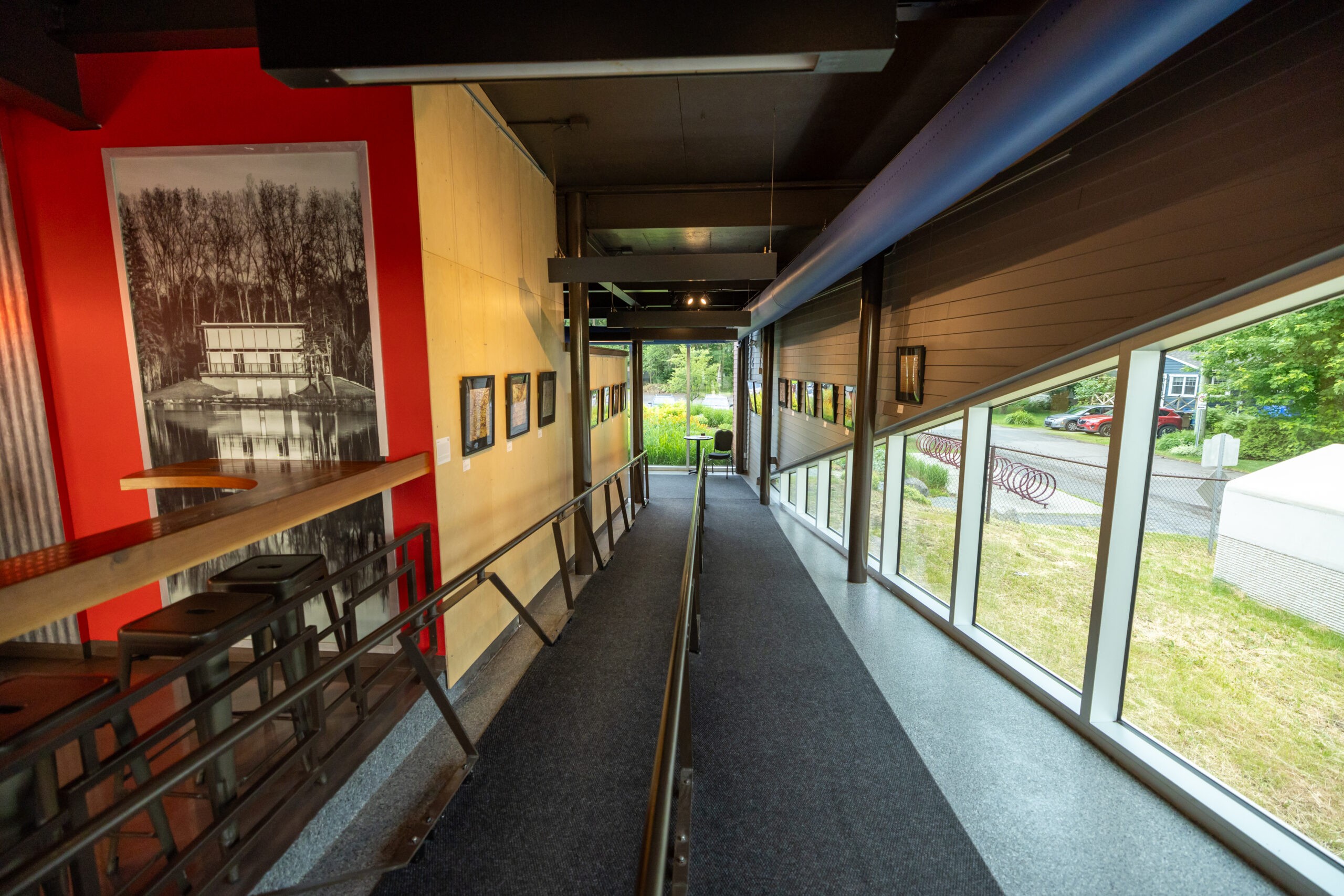 Une rampe intérieure moderne avec des rambardes mène à de grandes fenêtres donnant sur de la verdure et une route. Il y a des tabourets de bar à un comptoir sur la gauche et des photos encadrées sur les murs rouge et beige.