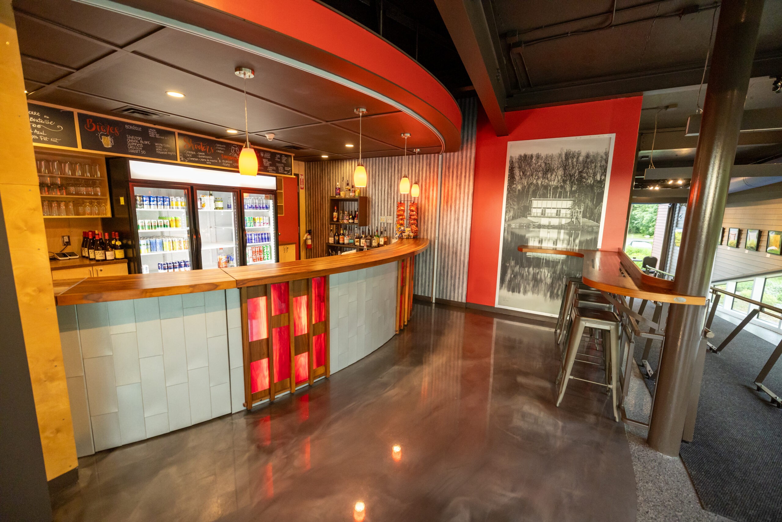 Un bar moderne avec un comptoir en bois incurvé, des panneaux rouges et blancs, des tabourets, un menu sur tableau noir, des réfrigérateurs avec des boissons et une grande photo en noir et blanc sur un mur rouge. La lumière naturelle pénètre par de grandes fenêtres.
