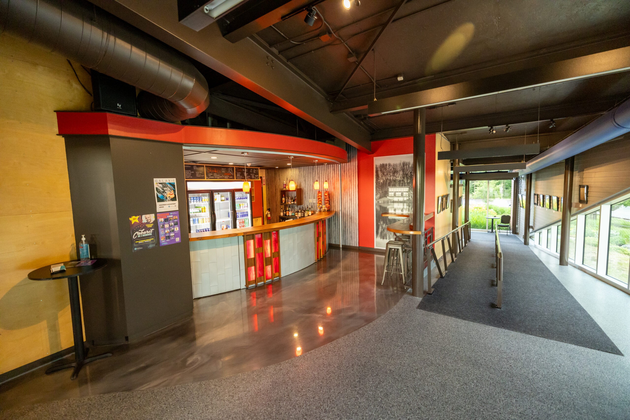 Un stand moderne avec un comptoir incurvé, des distributeurs de boissons et des snacks, situé dans un hall avec des affiches au mur et de grandes fenêtres laissant entrer la lumière naturelle. Une rampe d'accès mène à la sortie.