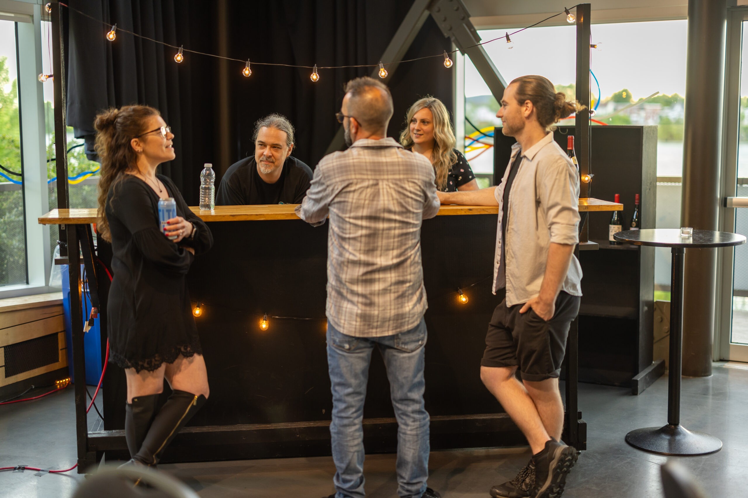 Cinq personnes sont rassemblées autour d'un bar en bois orné de guirlandes lumineuses ; deux sont derrière le bar, trois sont devant et discutent en toute décontraction. Le cadre est intérieur, avec de grandes fenêtres et une atmosphère joyeuse et détendue.