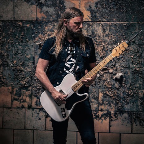 Un homme aux longs cheveux blonds joue de la guitare électrique devant un mur texturé et abîmé par les intempéries. Il porte une chemise noire et un jean, et semble concentré sur sa musique.