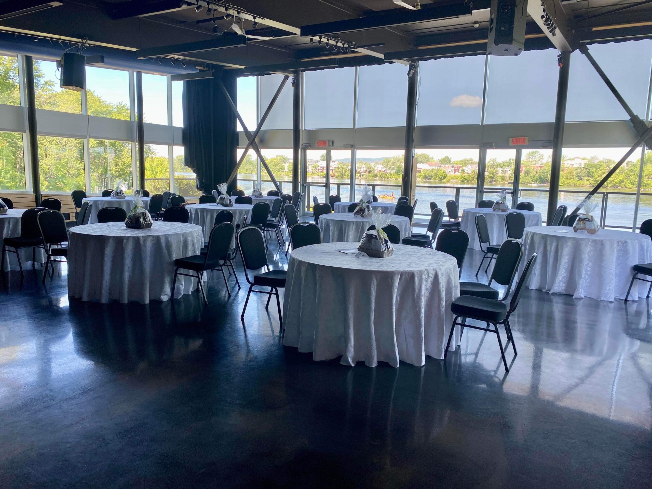 Une salle spacieuse avec des tables rondes recouvertes de nappes blanches et entourées de chaises, placées à côté de grandes fenêtres offrant une vue sur les arbres et l'eau à l'extérieur.