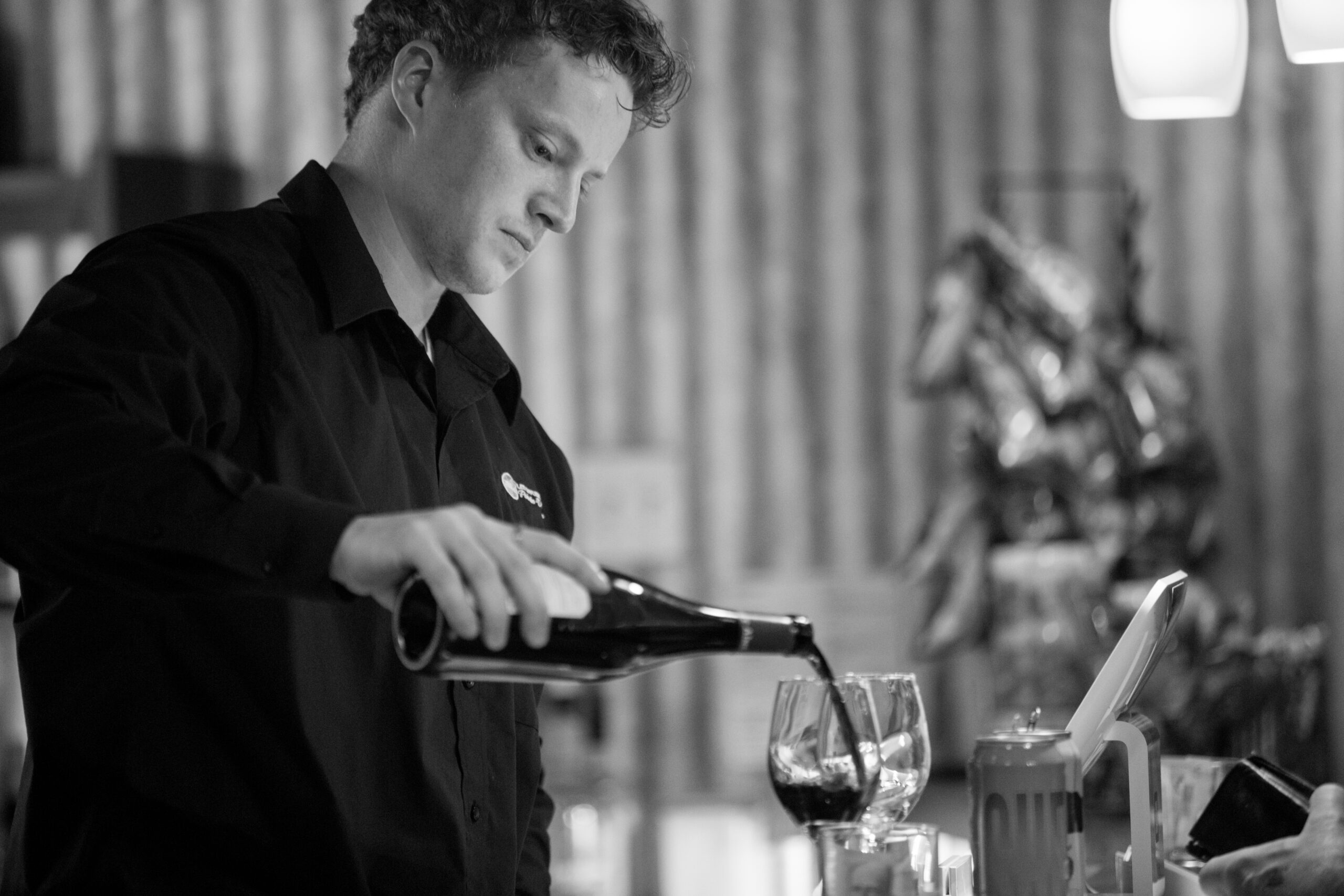 Un barman vêtu d'une chemise sombre verse du vin rouge dans un verre au bar, avec un autre verre et divers objets sur le comptoir, dans un décor intérieur légèrement éclairé. L'image est en noir et blanc.