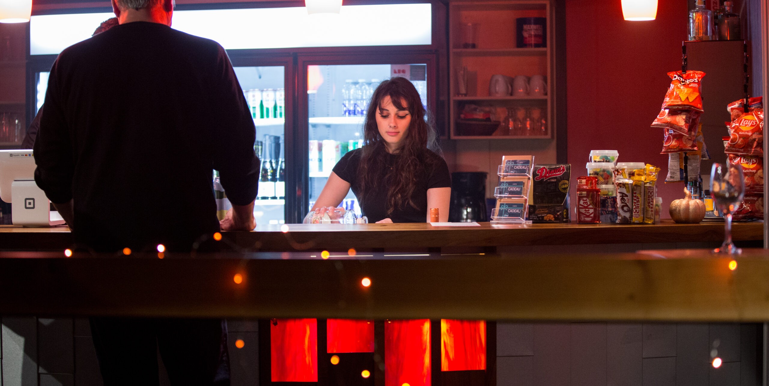 Une femme se tient derrière le comptoir d'un bar où sont présentés des snacks sur la droite. Un homme vêtu de sombre lui fait face de l'autre côté du comptoir. L'arrière-plan est éclairé par une lumière rouge et des étagères contenant des boissons et des verres.