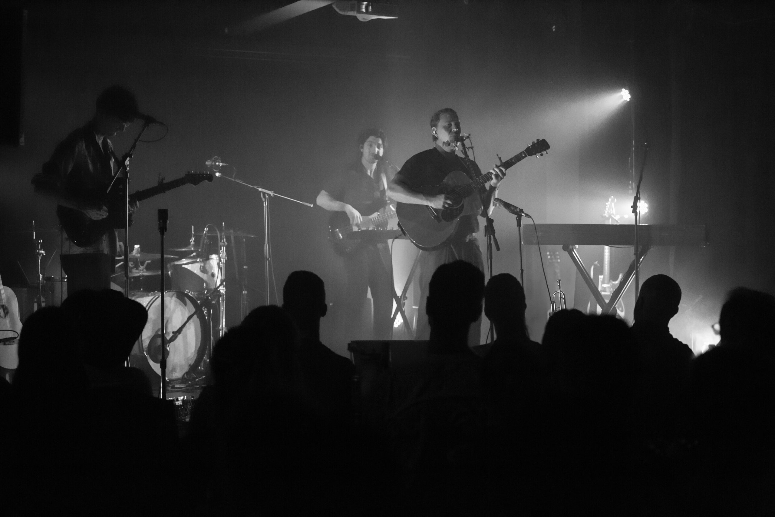 Photo en noir et blanc d'un groupe se produisant sur scène avec des guitares, une batterie et un clavier. Les membres du groupe sont éclairés par les lumières de la scène, tandis que le public apparaît en silhouette au premier plan.
