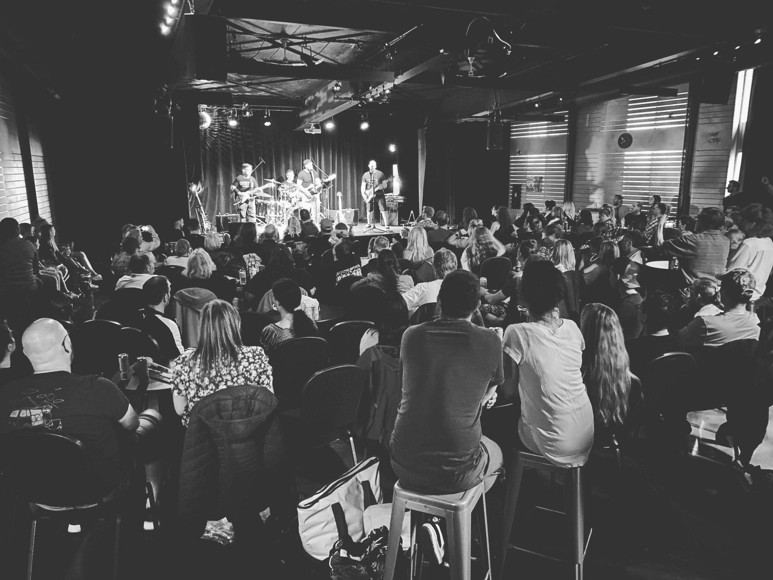 Cette photo en noir et blanc d'une salle de spectacle bondée montre des personnes assises qui regardent un groupe se produire sur scène sous les projecteurs. Le public est tourné vers l'avant et l'atmosphère semble animée et engagée.
