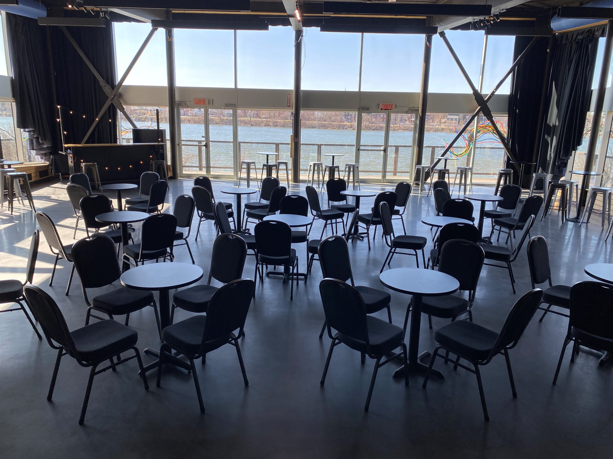 Une salle spacieuse avec des tables rondes et des chaises noires, disposées en rangées. De grandes fenêtres laissent entrer la lumière du soleil et offrent une vue sur la rivière et les arbres à l'extérieur. Un petit bar se trouve dans le coin gauche, et des tables hautes sont disposées le long des fenêtres.