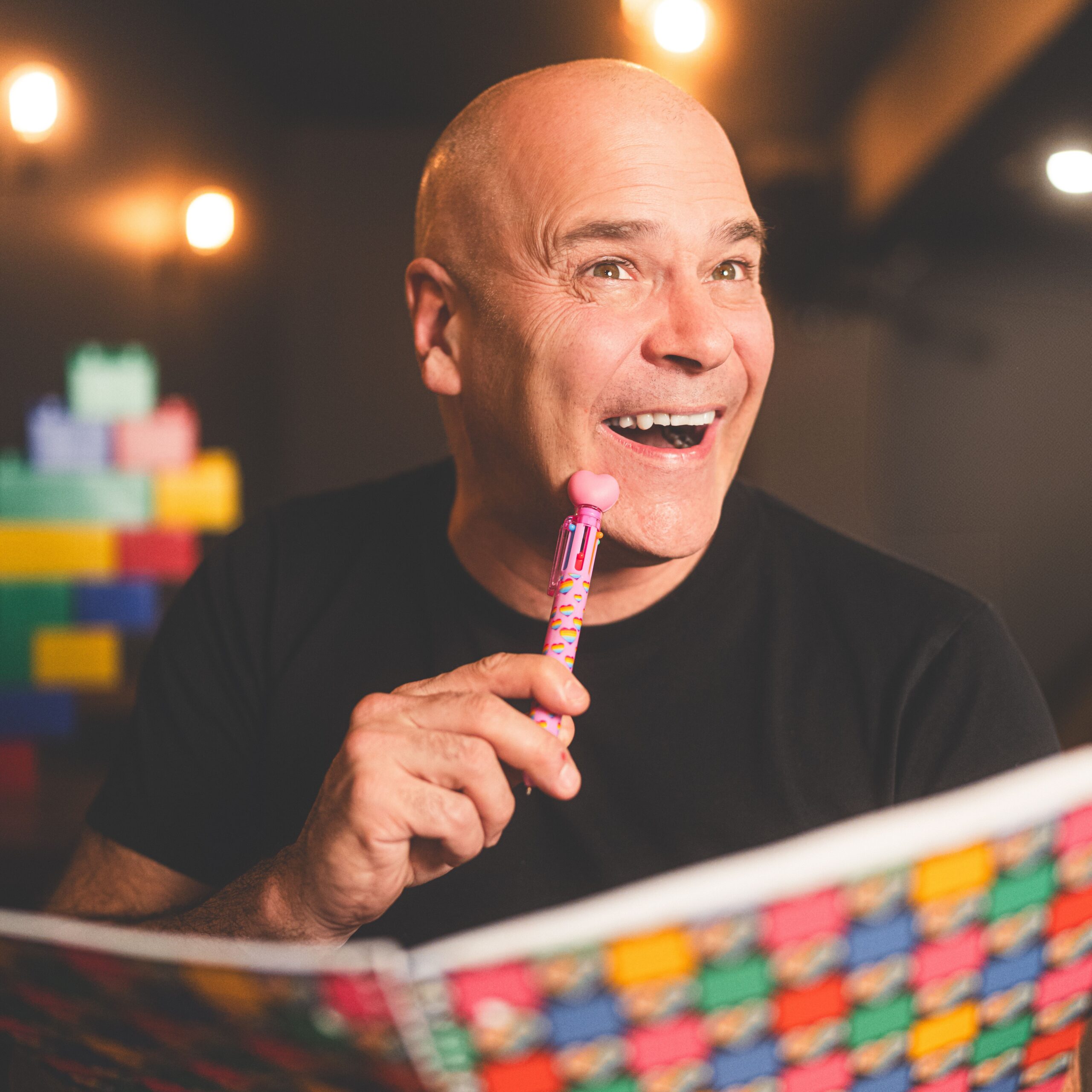 Un homme chauve souriant, vêtu d'une chemise noire, porte un stylo coloré à son menton et regarde avec enthousiasme une grande feuille colorée. Des blocs de jouets flous et surdimensionnés se trouvent à l'arrière-plan et bénéficient d'un éclairage chaud.