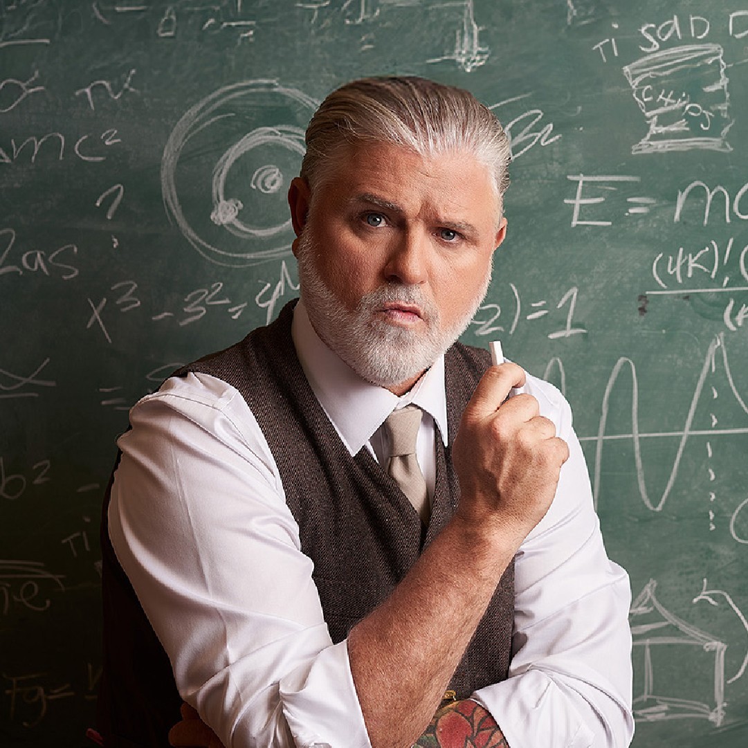 Un homme âgé, sérieux, aux cheveux et à la barbe blancs, vêtu d'un gilet et d'une cravate, tient une craie devant un tableau couvert d'équations et de diagrammes mathématiques.