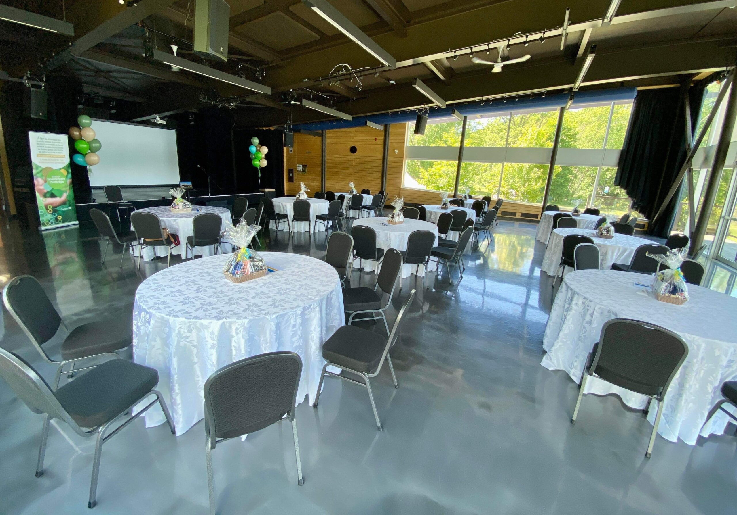 Une salle spacieuse avec des tables rondes recouvertes de tissus blancs, chacune avec un petit centre de table, des chaises noires, un écran de projection et des colonnes de ballons verts et blancs près de grandes fenêtres laissant entrer la lumière naturelle.