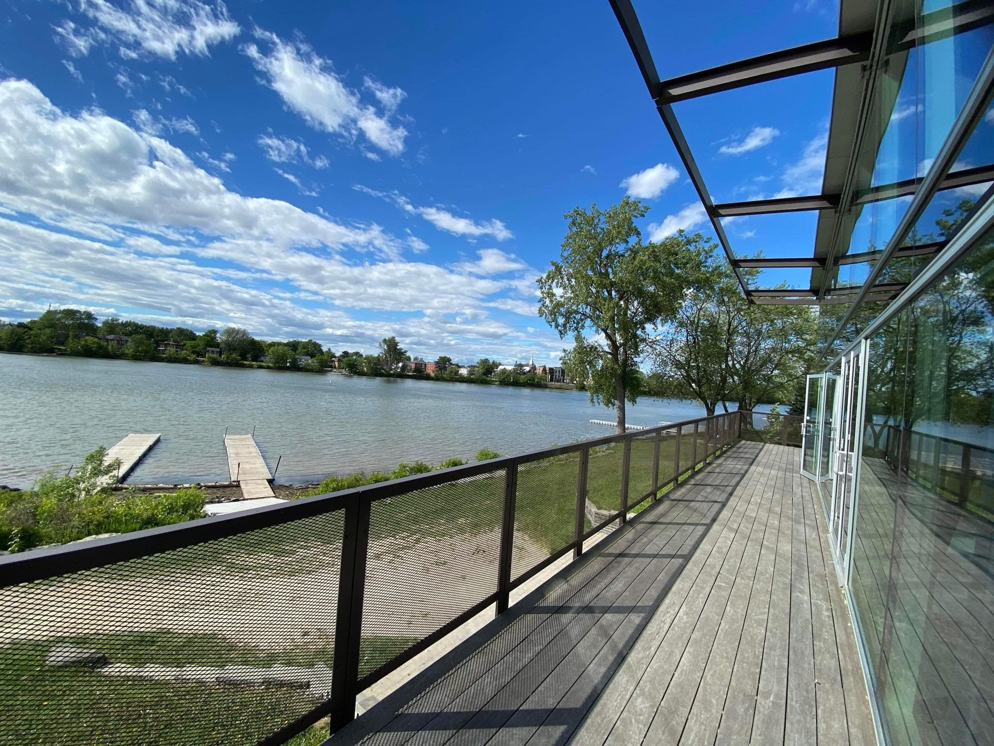 Une terrasse en bois avec une rambarde en métal surplombe une rivière calme, avec un quai s'avançant dans l'eau. Des arbres et de la verdure bordent la rive opposée sous un ciel bleu parsemé de nuages.