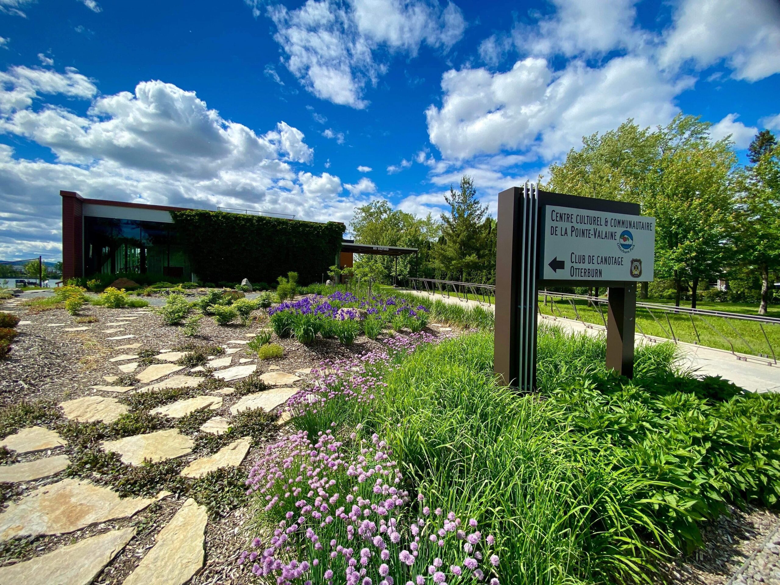 Un chemin de pierre serpente à travers un jardin vibrant de plantes violettes et vertes, menant à un bâtiment moderne. Un panneau indique "Centre Culturel & Communautaire de la Pointe-Valaine" et pointe vers la gauche sous un ciel partiellement nuageux.