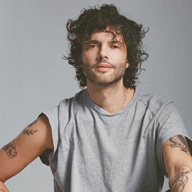 Un homme aux cheveux noirs bouclés et tatoués sur les deux bras porte un t-shirt gris clair et regarde l'appareil photo avec une expression neutre sur un fond clair uni.