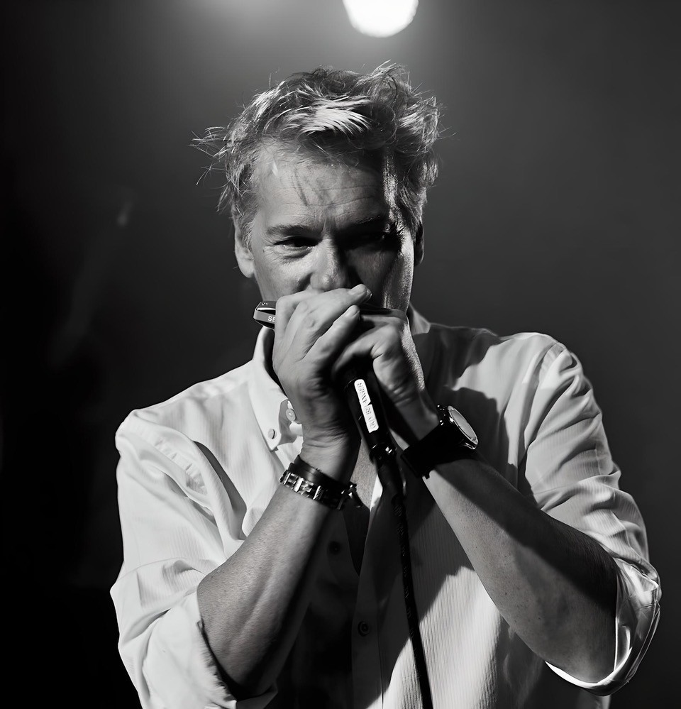 Un homme aux cheveux ébouriffés joue de l'harmonica et chante dans un micro sur scène, vêtu d'une chemise blanche boutonnée ; un éclairage dramatique crée une atmosphère sombre, en noir et blanc.
