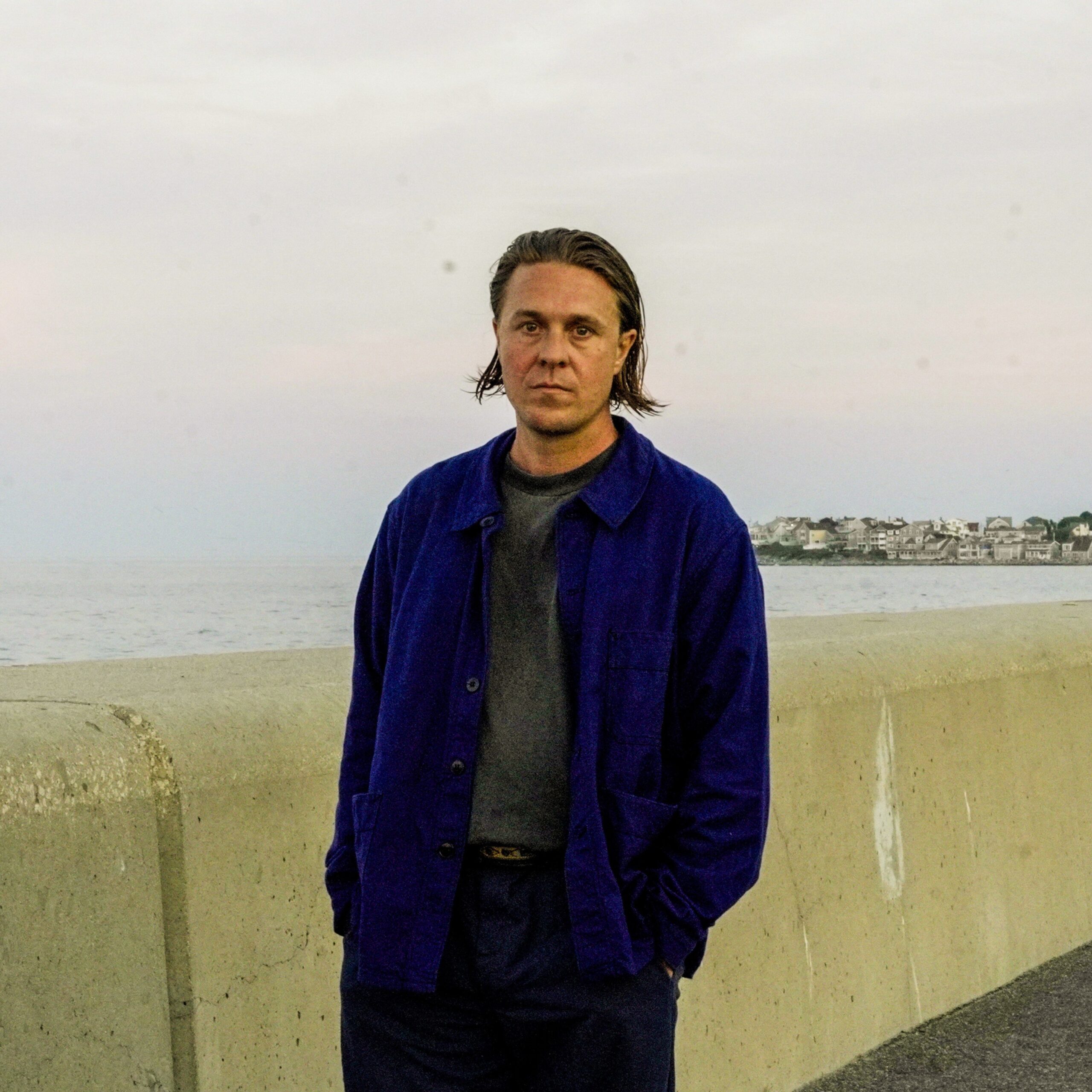 Une personne aux cheveux mi-longs se tient près d'une digue en béton, vêtue d'une veste bleue et d'un pantalon sombre. L'océan et un rivage lointain avec des bâtiments sont visibles à l'arrière-plan sous un ciel pastel.